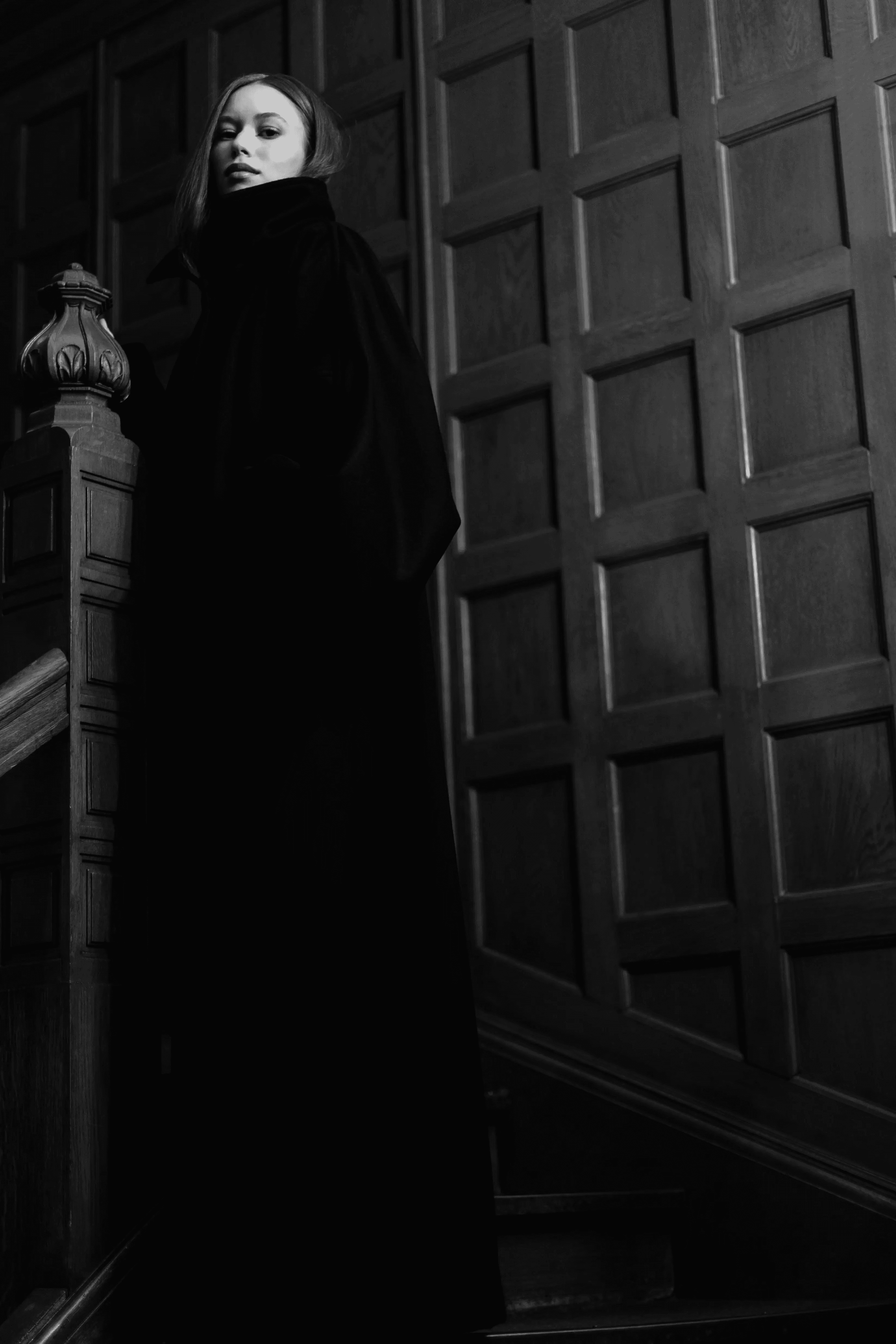 Black and white fashion image of a woman posing by a door in a dark coat.