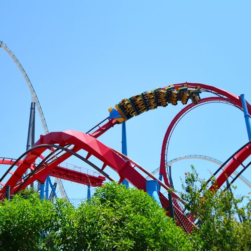 A vibrant red rollercoaster with multiple loops and cars full of people riding, set against a clear blue sky and green foliage.