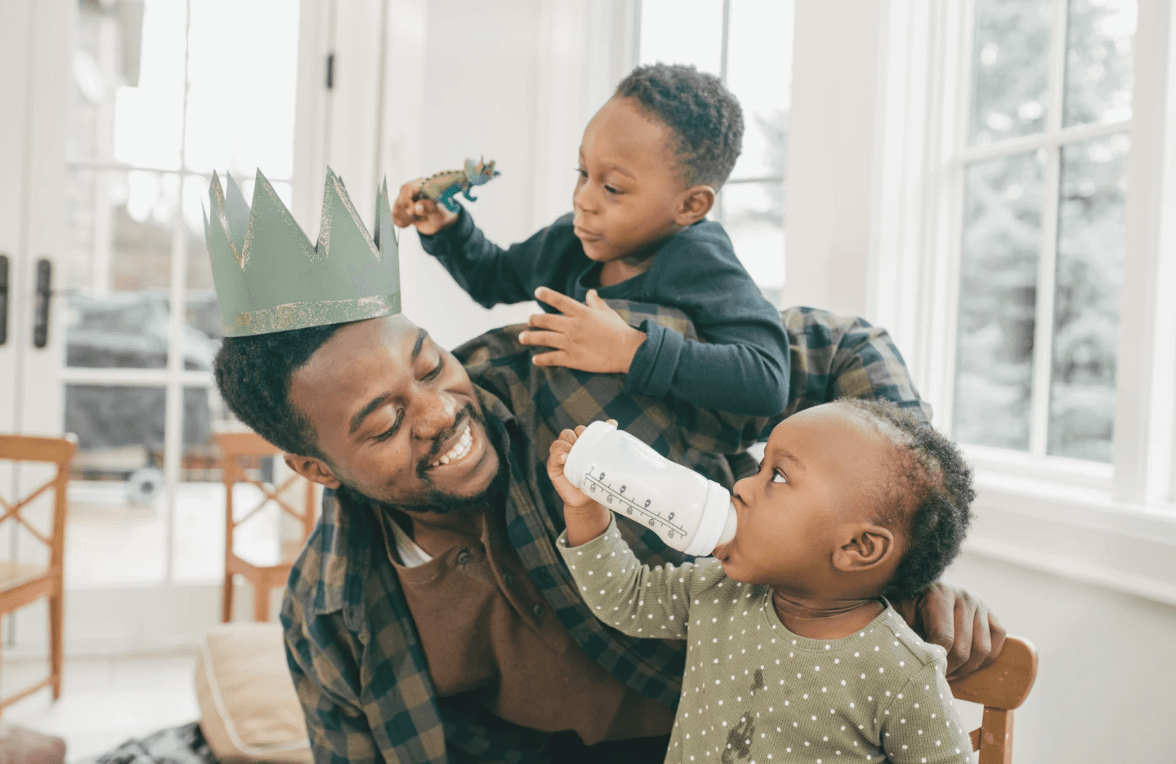 A joyful dad plays with his two young children, one on his back and the other sitting, both smiling and engaged.