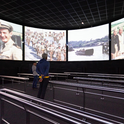 Zwei Personen stehen vor vier großen Bildschirmen, die historische Militäraufnahmen in einem dunklen Raum mit Metallgeländern zeigen.