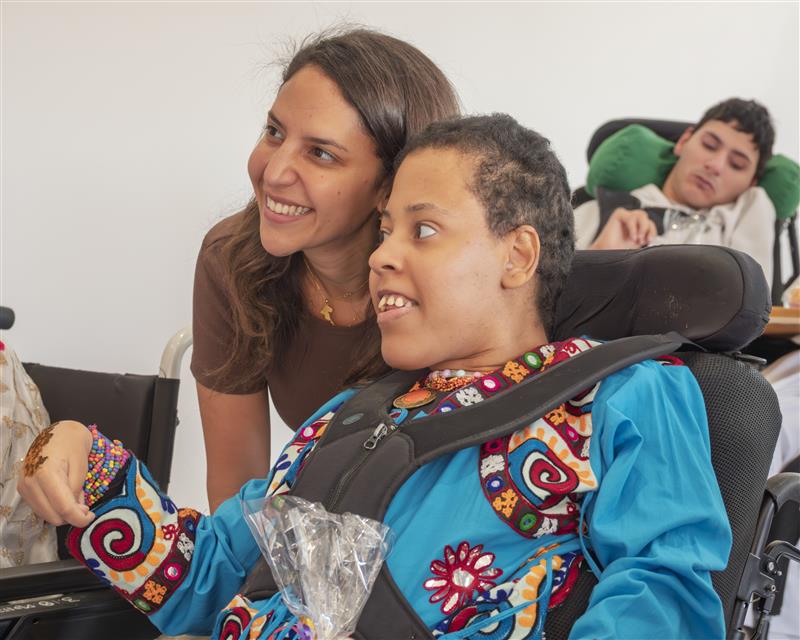 Una persona con una camiseta negra y pantalones de color claro está de pie en la cima rocosa de una montaña al atardecer, sosteniendo una cámara frente a su cara para tomar una foto. Un mar de nubes se extiende detrás de ellos, iluminado por la luz dorada del sol poniente.