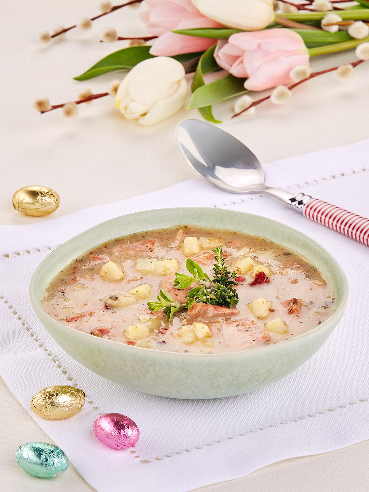 A pastel green bowl of creamy soup zurek garnished with fresh herbs on a white napkin, surrounded by decorative Easter eggs and pink tulips, showcasing a delightful food photo session.