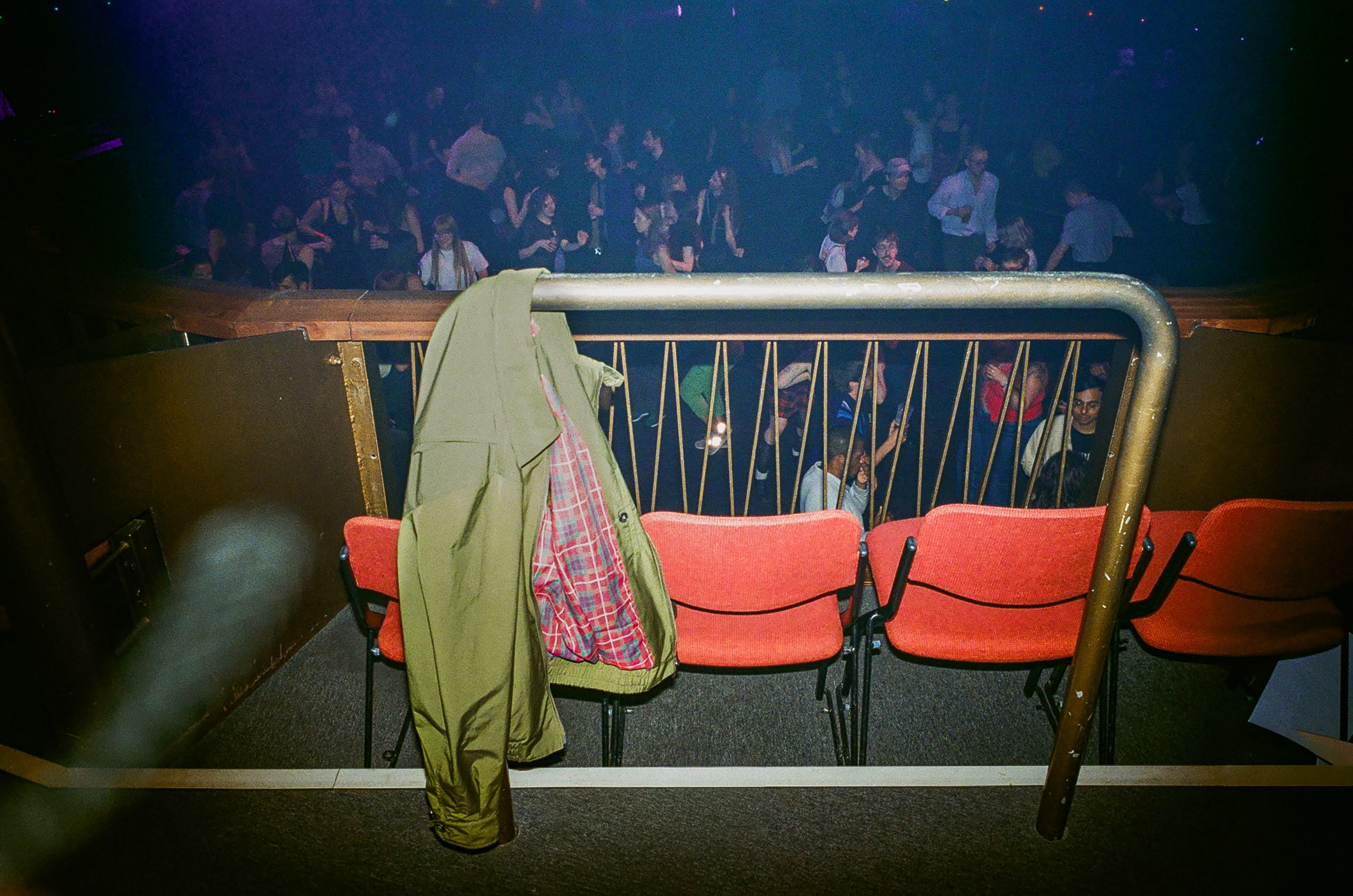 Dans un night club, un blouson de pluie vert et doublure tartan de la marque Baracuta est accroché derrière des fauteuils au dessus de la foule