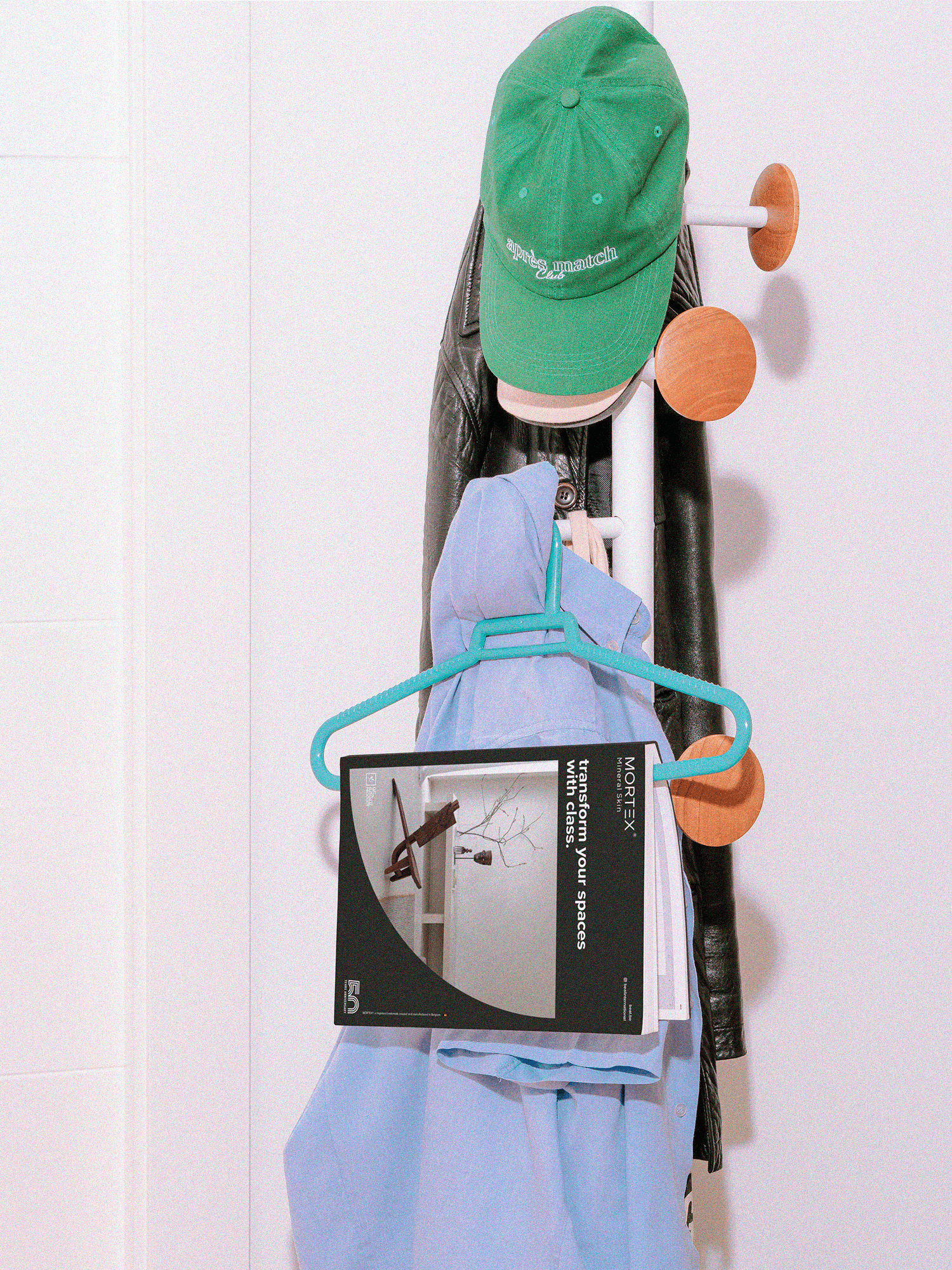 a casual hallway scene featuring a coat rack holding a green cap, a leather jacket, and a light blue shirt. A Mortex brochure hangs neatly from a turquoise hanger, adding a styled yet lived-in feel to the composition.