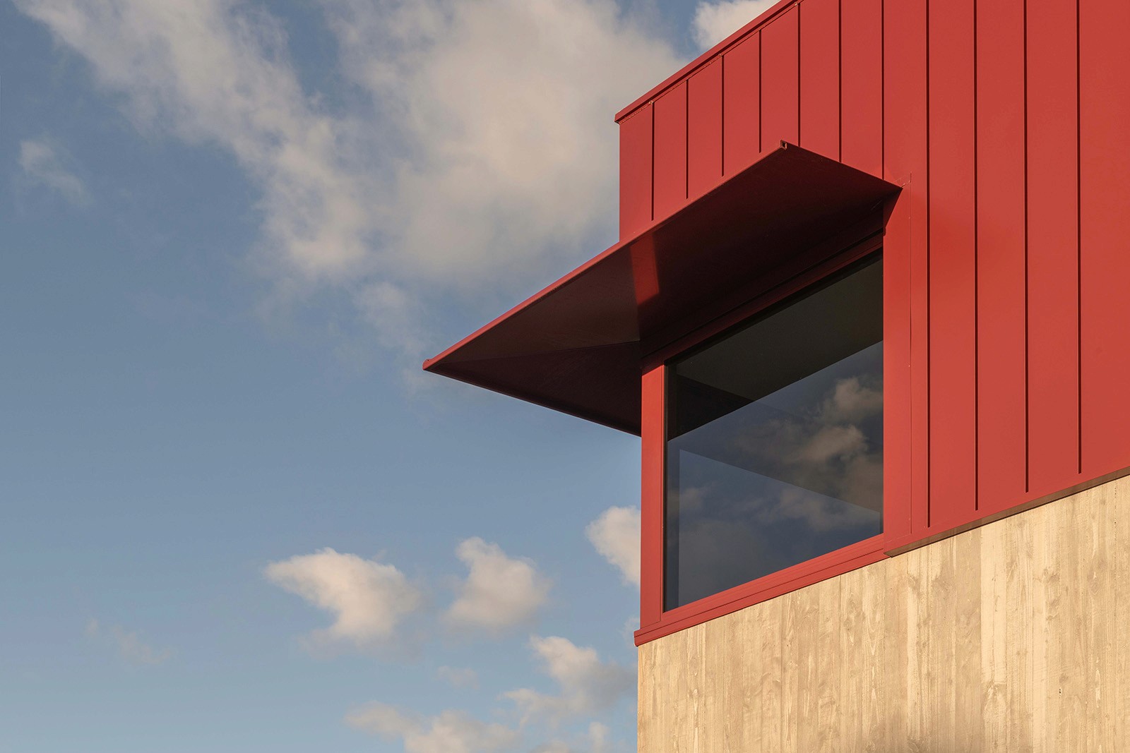 Architectural detail of the RedSheds project, showing a red metal-clad upper volume with a projecting canopy and glazed window set against a concrete base and open sky.