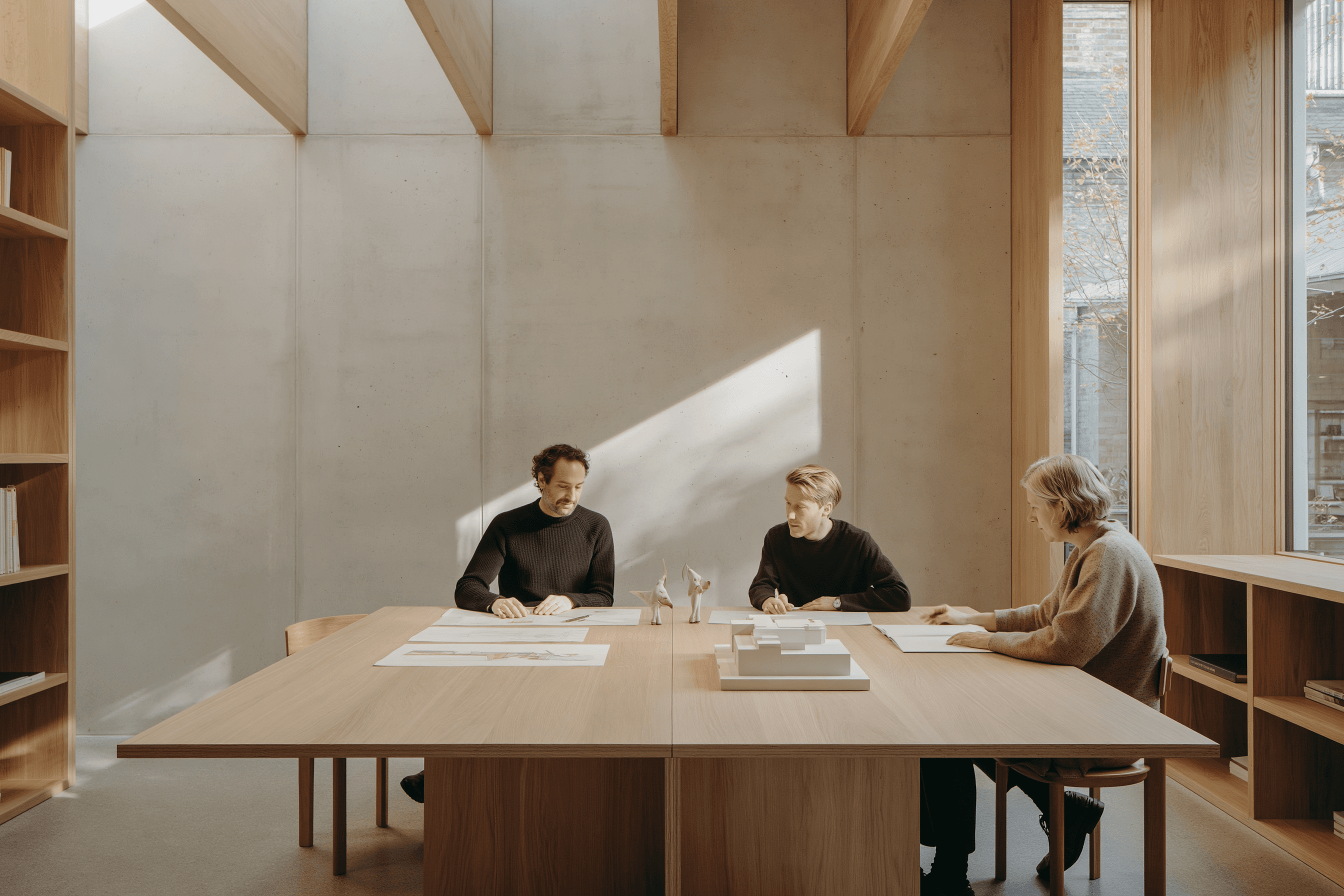Two architects in quiet conversation over a site plan in a darkened studio, warm amber light above the table