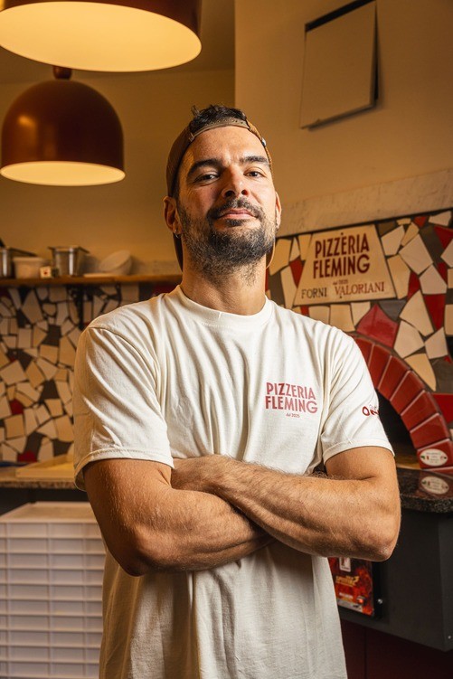 A man with crossed arms stands confidently in a café environment, with warm lighting and a casual atmosphere.