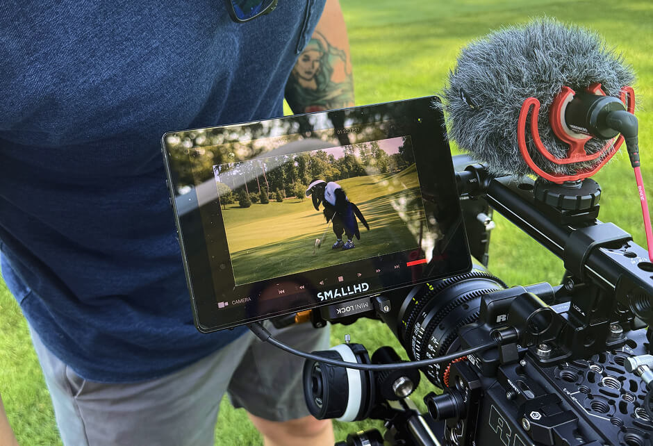 A person operates a professional video camera with a monitor displaying a golfer preparing to swing on a green golf course.