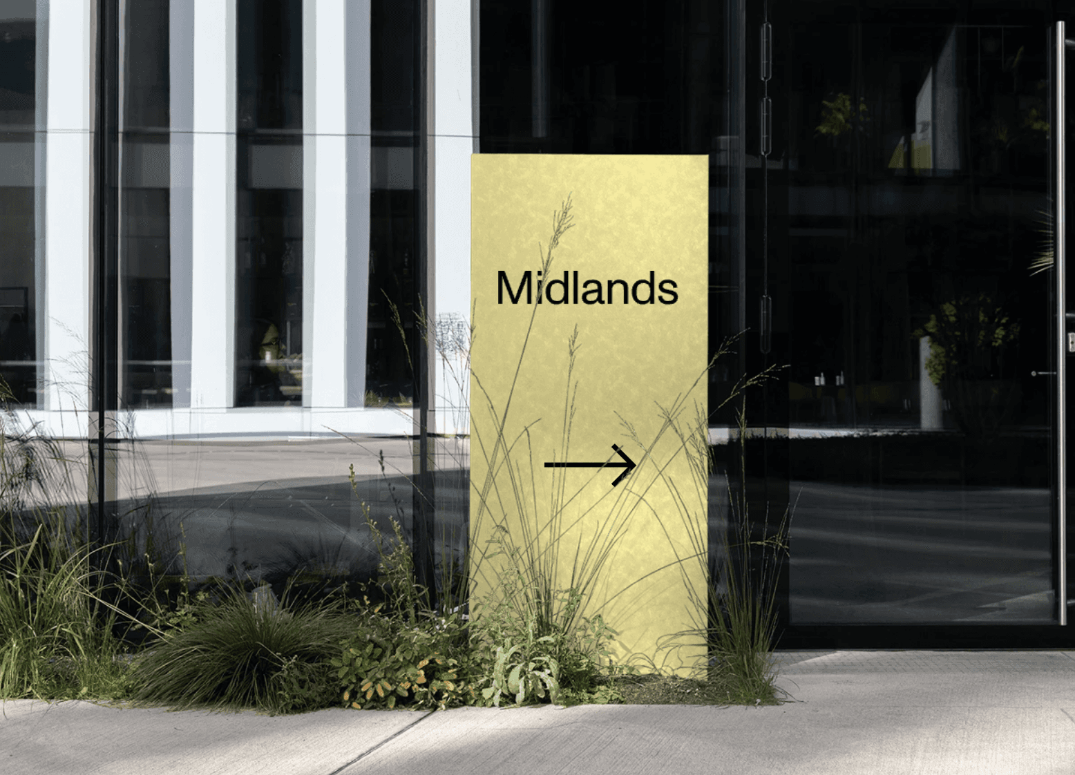 Yellow sign with "Midlands" text and arrow, surrounded by tall grass, pointing towards building entrance.