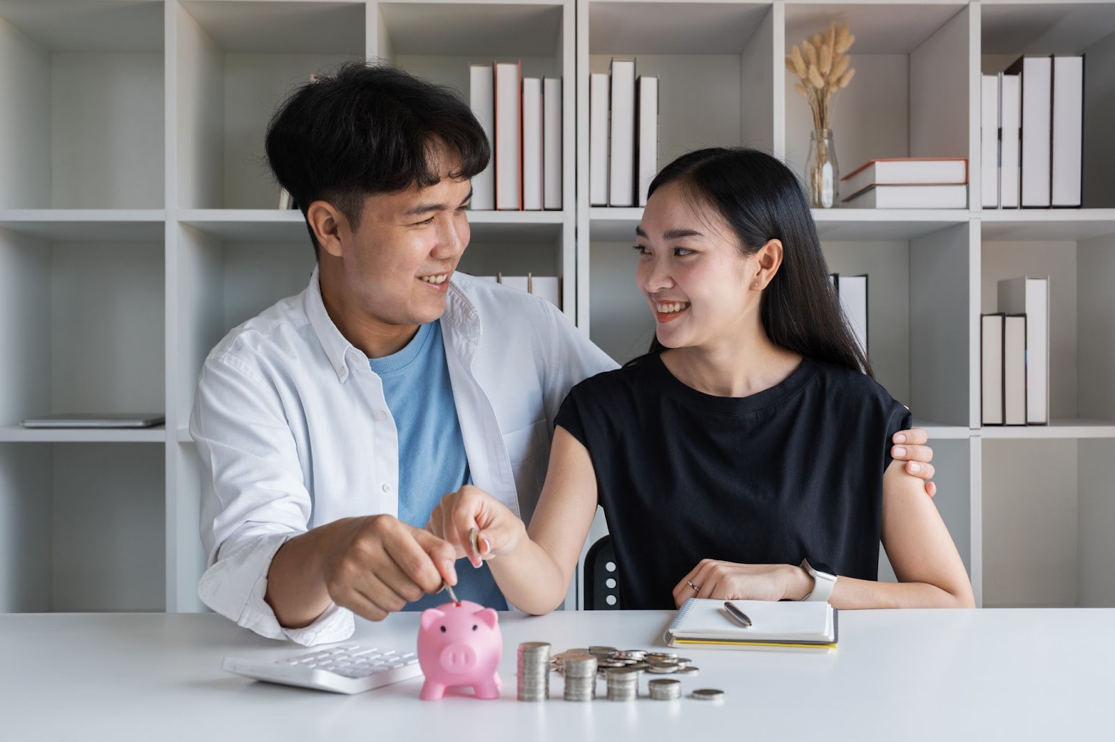 Couple adding money to a piggy bank with a bookshelf in the background. 