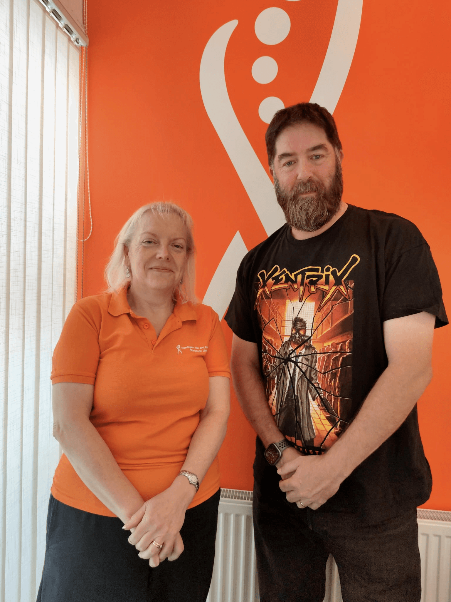 FTwo people stand in front of an orange wall. A woman on the left wears an orange polo shirt. A man on the right wears a black t-shirt with a graphic design.eln t em etet