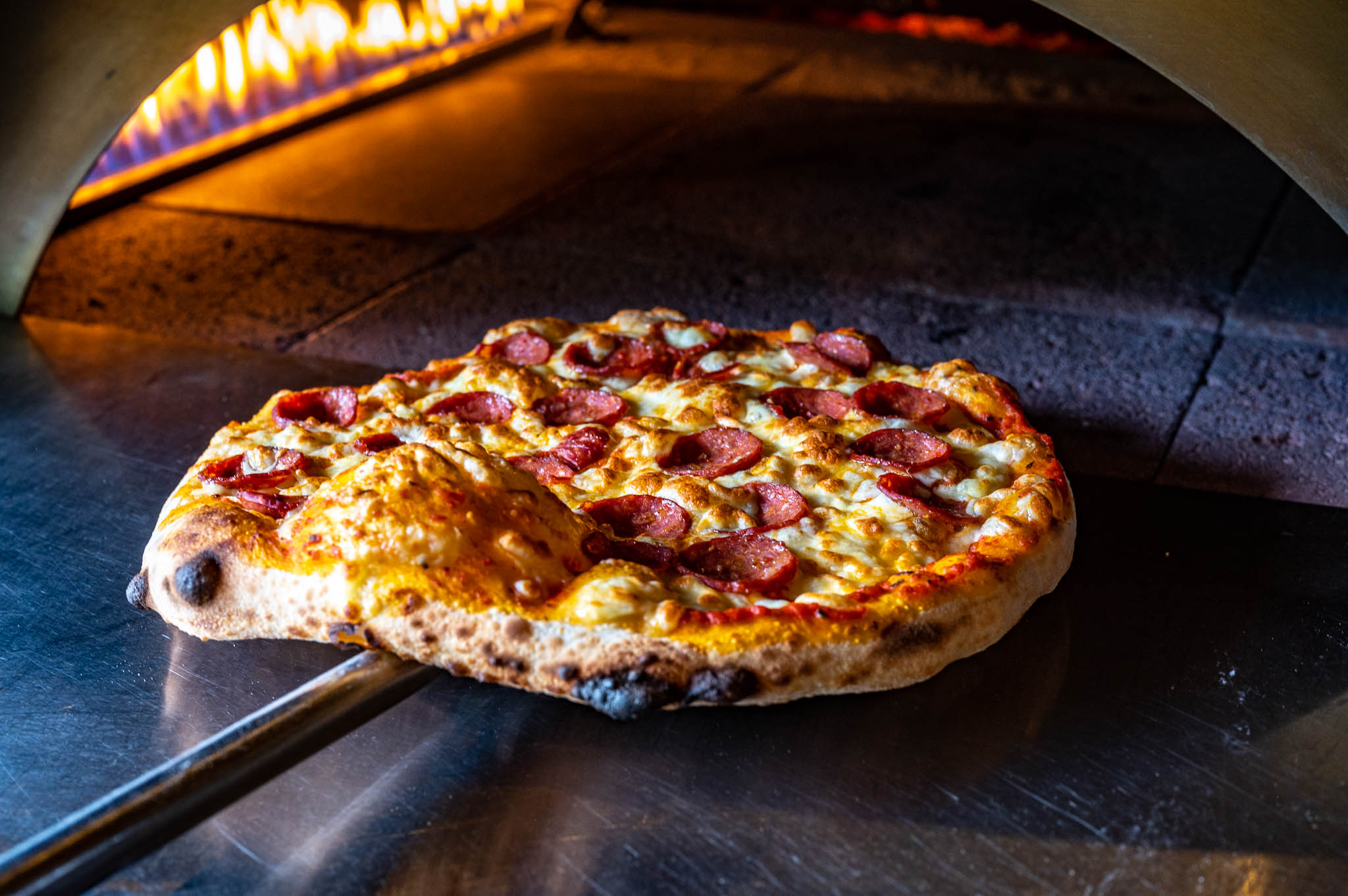 Freshly baked pepperoni pizza being taken out of the wood-fired oven, topped with arugula.