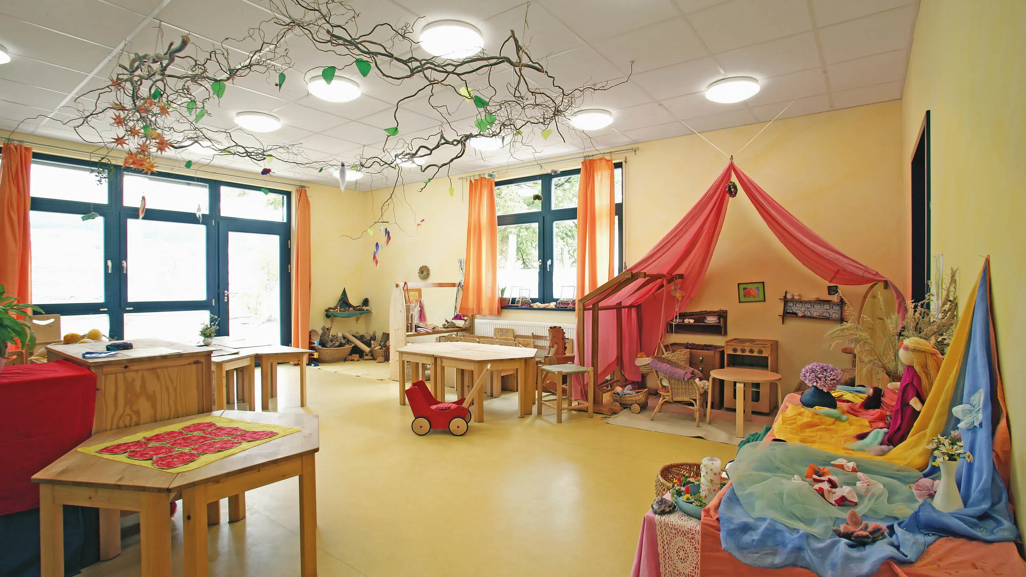 Ein heller Gruppenraum mit Holztischen, Spielzeug, einem Spielzelt aus roten Stoffen und dekorierten Ästen an der Decke.