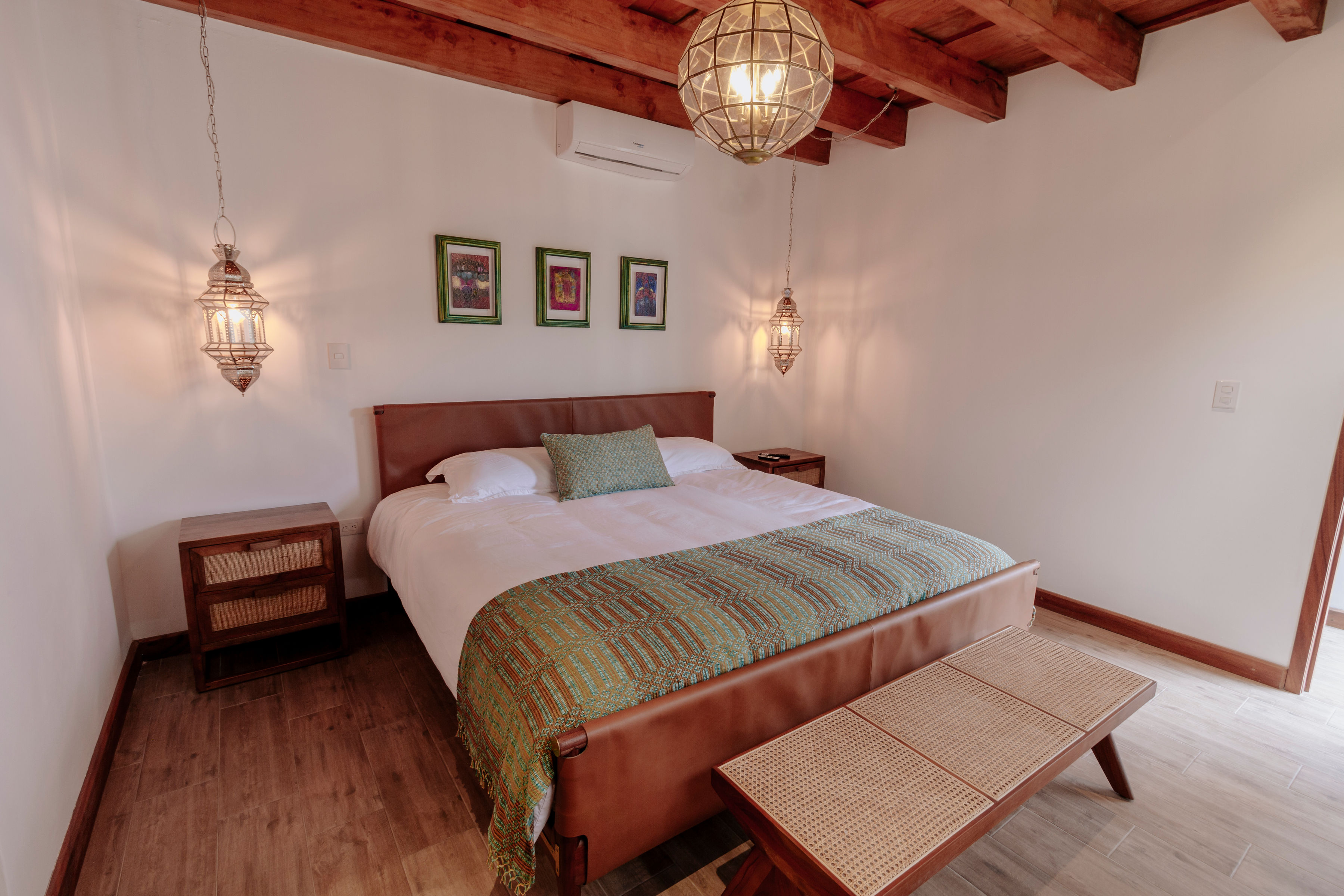 A cozy bedroom with a wooden beamed ceiling features a large double bed adorned with a green patterned throw, flanked by two pendant lights and framed artwork on the wall, complemented by rustic wooden furniture.
