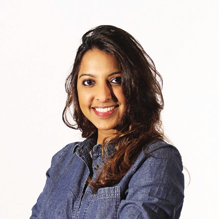 A woman with medium-length dark brown hair with lighter highlights smiles at the camera. She wears a blue denim jacket over a white collared shirt. The background is plain white.