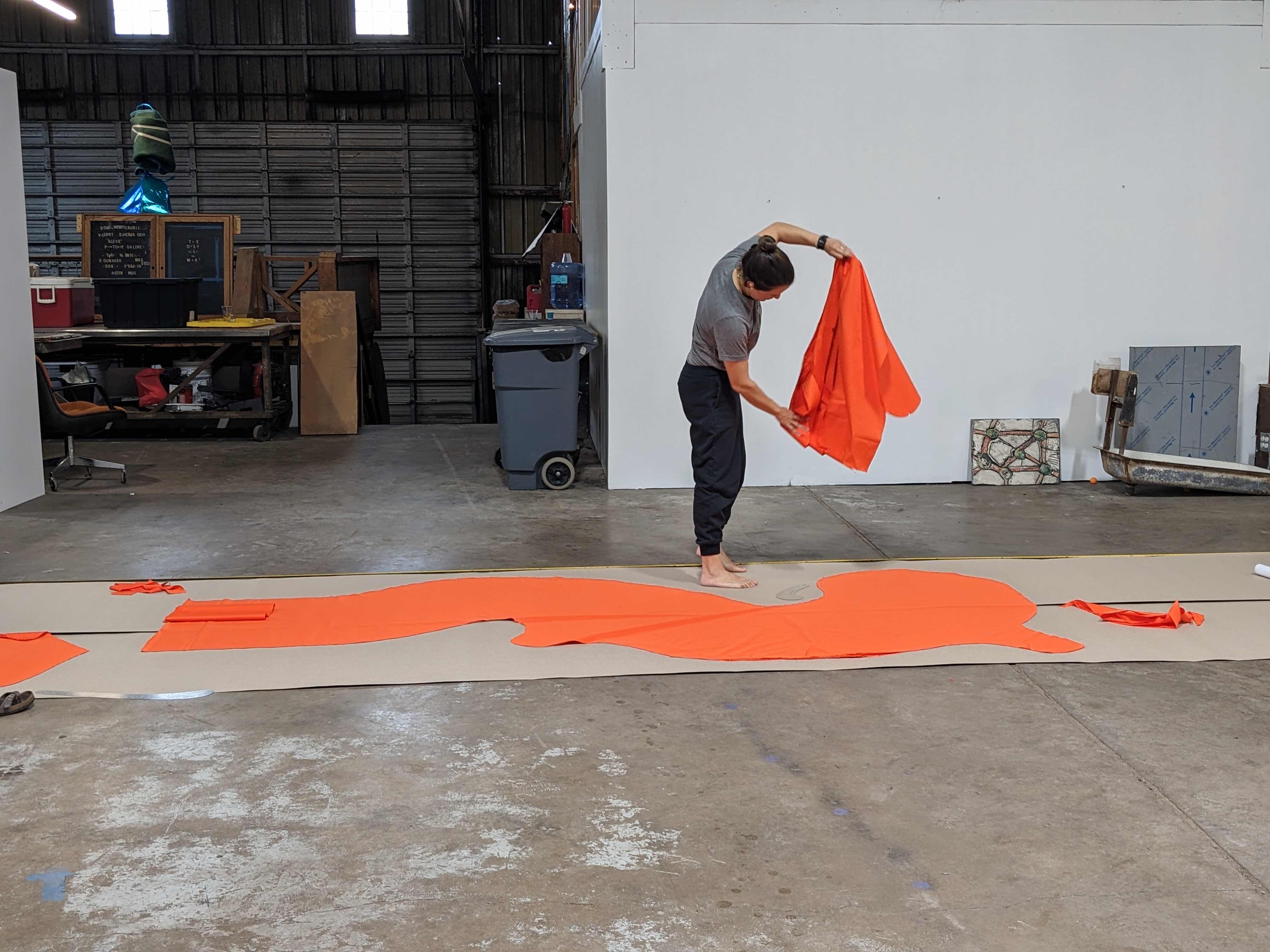 A person unfolds an orange tarp on a concrete floor in a large, open space.
