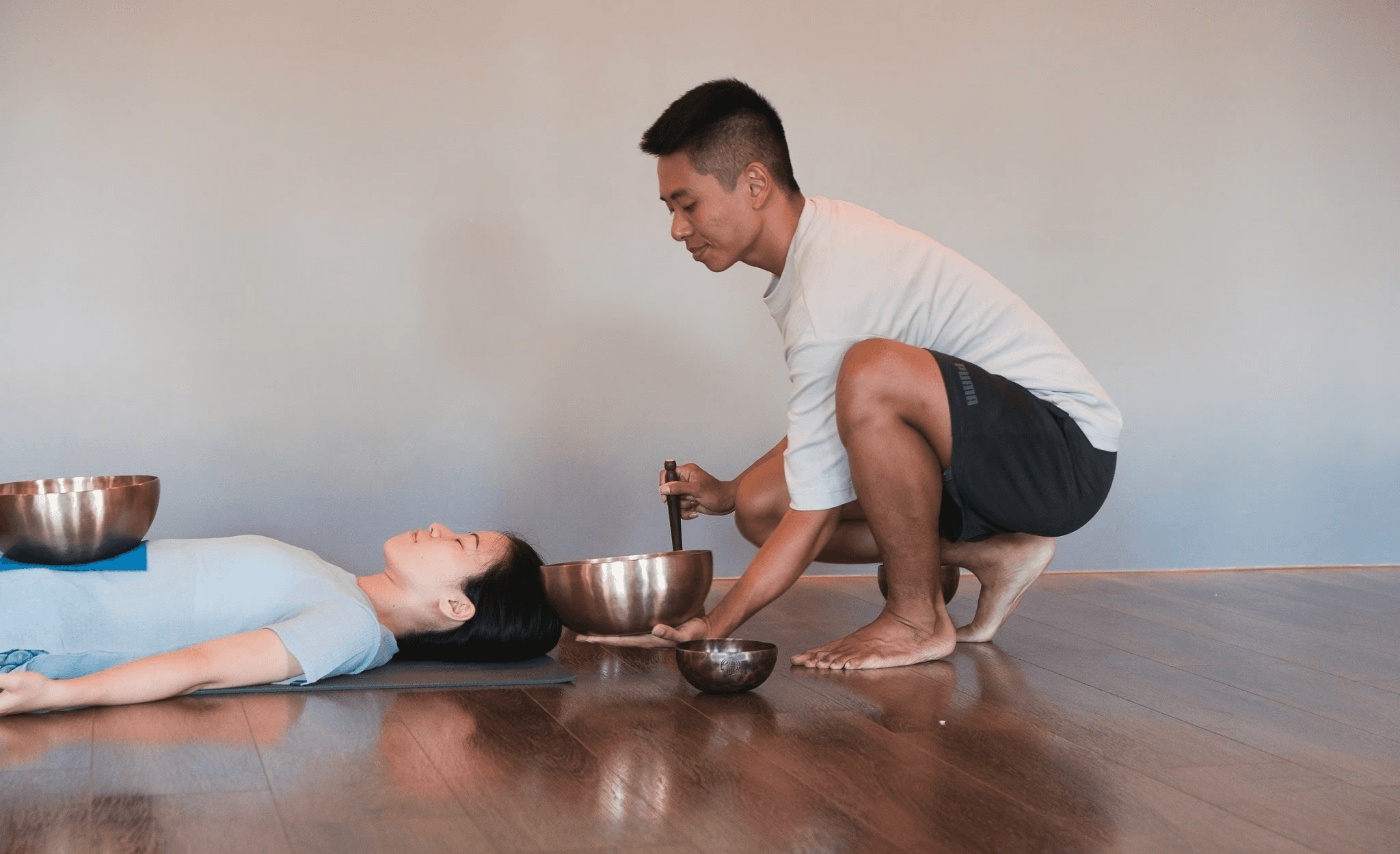 在一個寧靜的房間裡,一名男引導師蹲在一名躺在瑜珈墊上的女子身旁,在她的頭旁拿著一個頌缽,而另一個碗則放在她的胸前,房間裡的地板是木質的,營造出一種安全感受的氛圍。