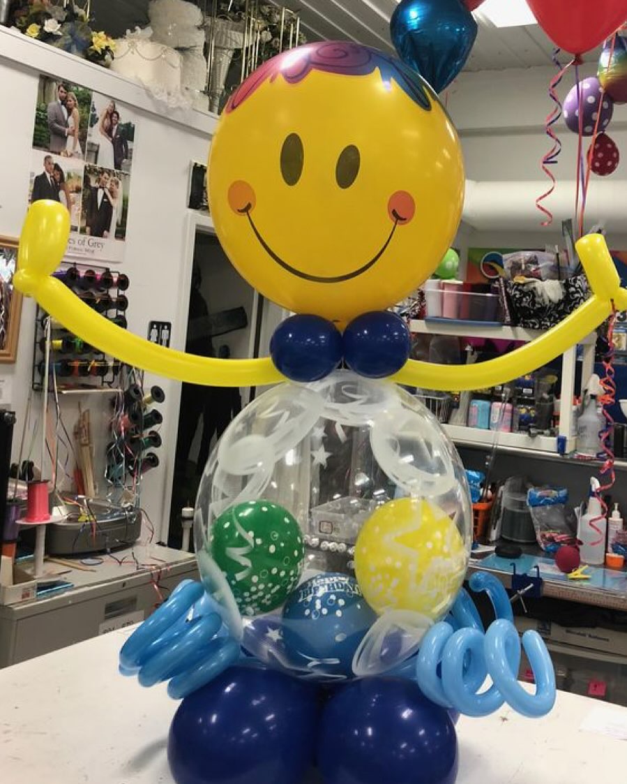 Balloon sculpture featuring a large yellow smiley face with colorful balloon accents and outstretched arms made by Be Bold Balloons