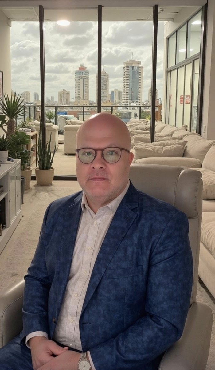 Man wearing glasses and a blue blazer sitting indoors with a city skyline visible through large windows in the background.