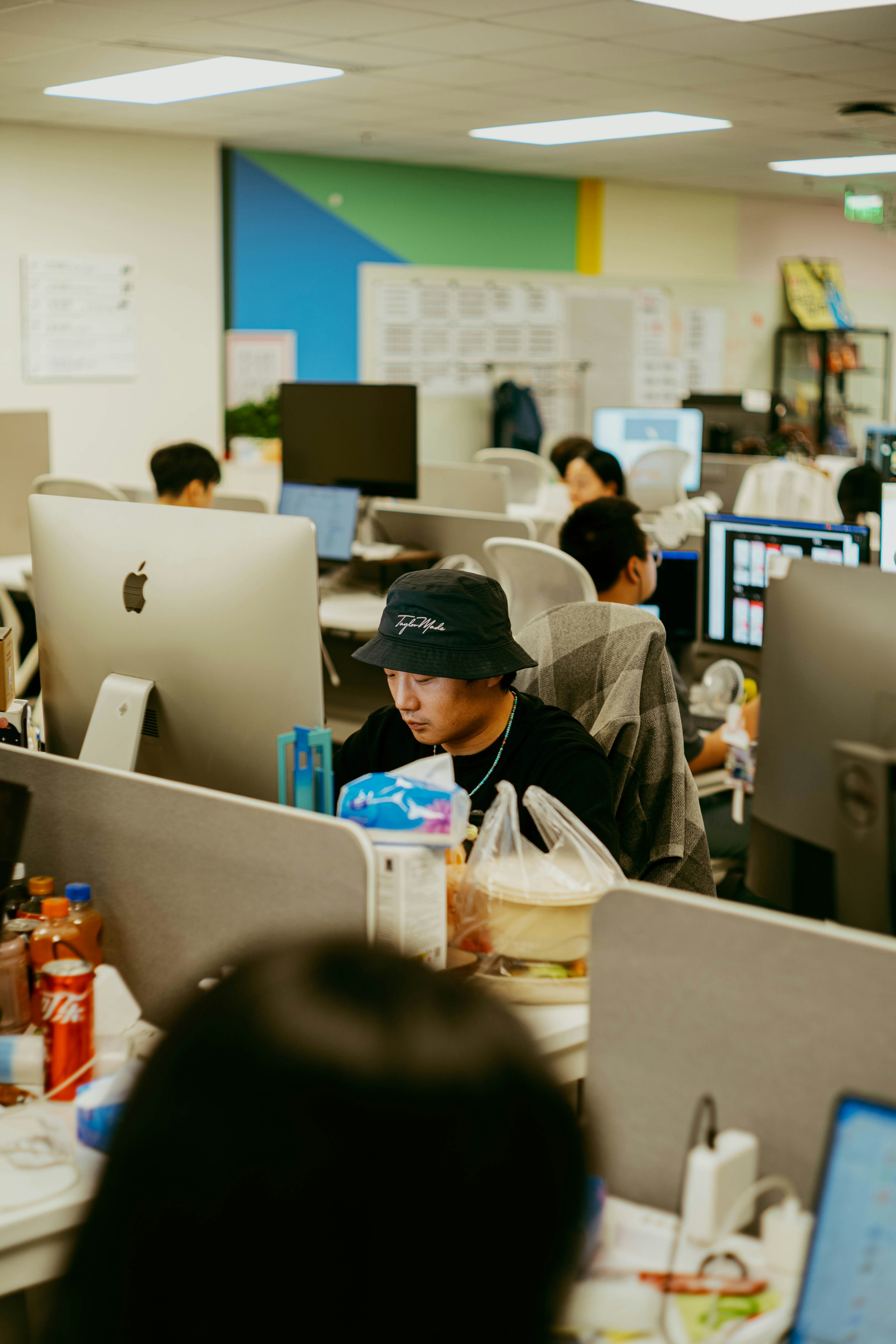 An office scene with several people working at desks, focused on screens, with light-colored walls and casual decor.