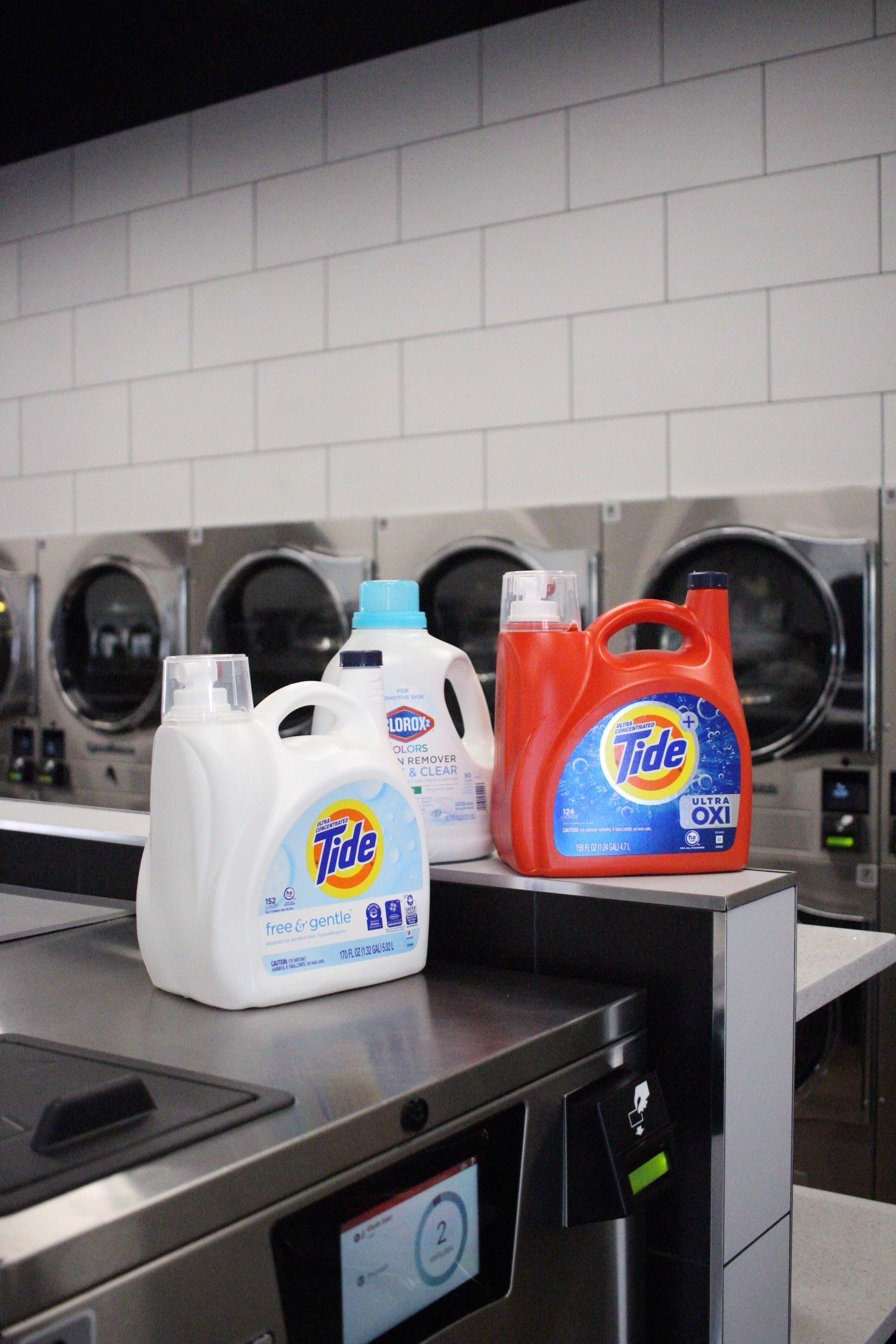 Laundry detergent bottles on a folding table in front of stainless steel washers, highlighting operational supplies to factor into how to buy a laundromat.