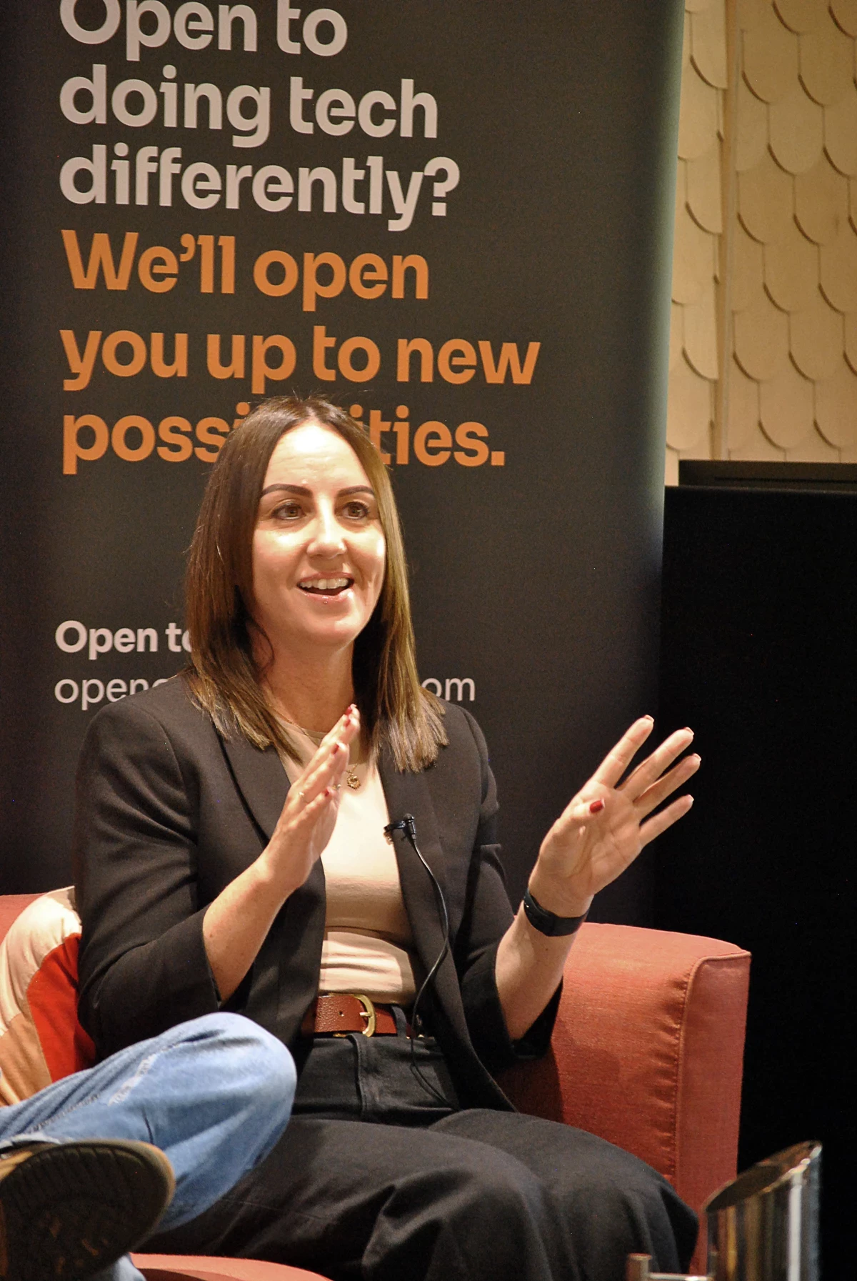 Person seated on a red chair speaking with hands raised, in front of a banner that reads “Open to doing tech differently? We’ll open you up to new possibilities.”