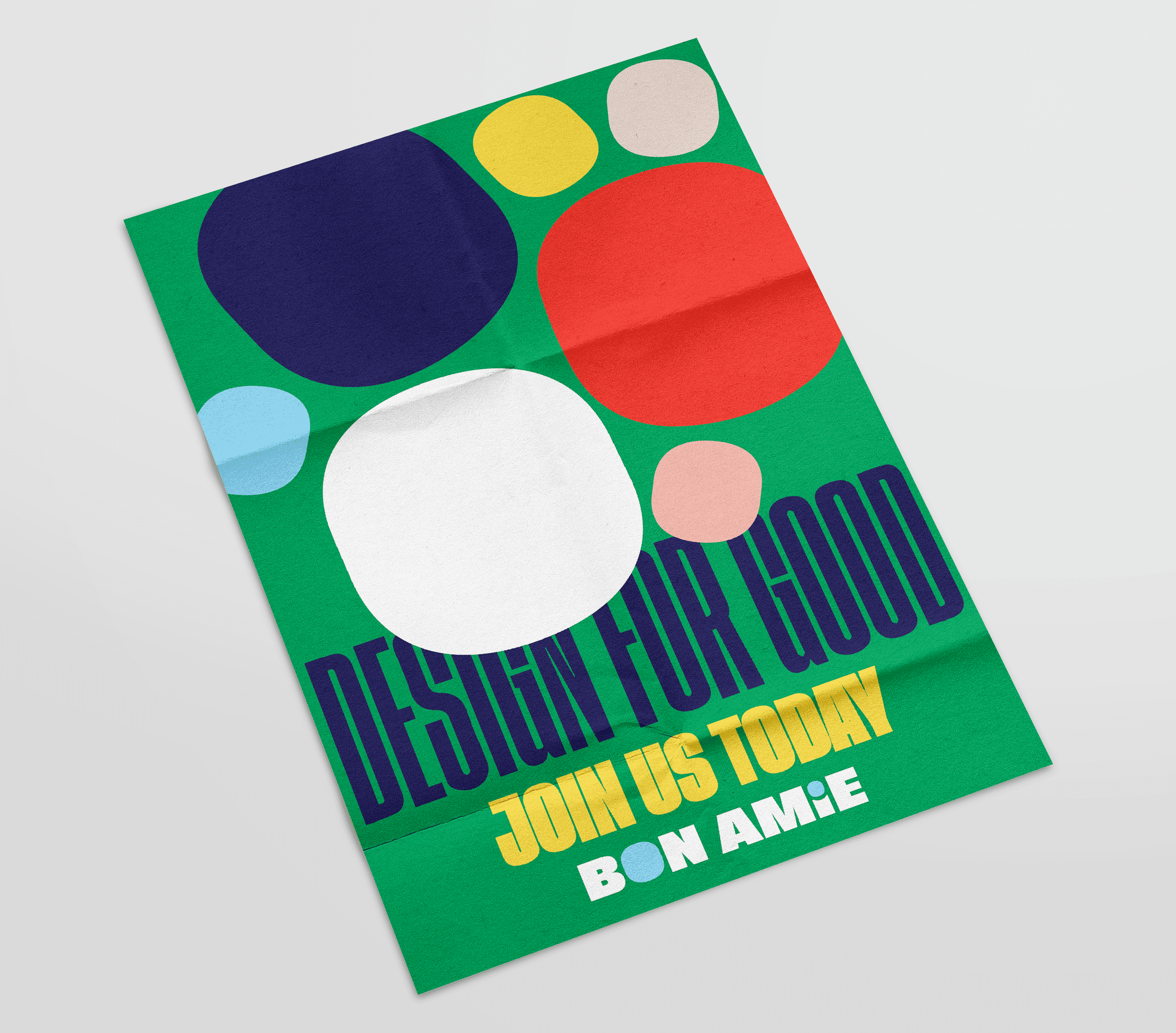 A green rectangular poster with rounded corners displays a collection of overlapping circles in blue, red, yellow, and white. The text "DESIGN FOR GOOD" is printed in dark blue, with "JOIN US TODAY" in yellow and "BON AMIE" in white below it. The poster is angled slightly to the right.