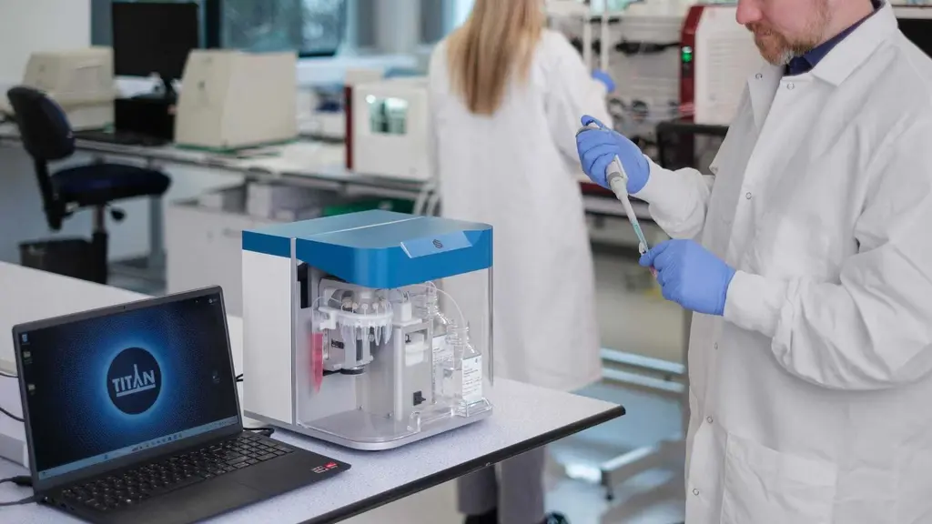 Man in a white lab coat and blue gloves using a pipette beside a blue and white Titan bench instrument, with a laptop showing the Titan logo