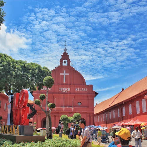 Tourists walking near Christ Church Melaka, a red building with a large white cross, in a square under a bright, blue sky.