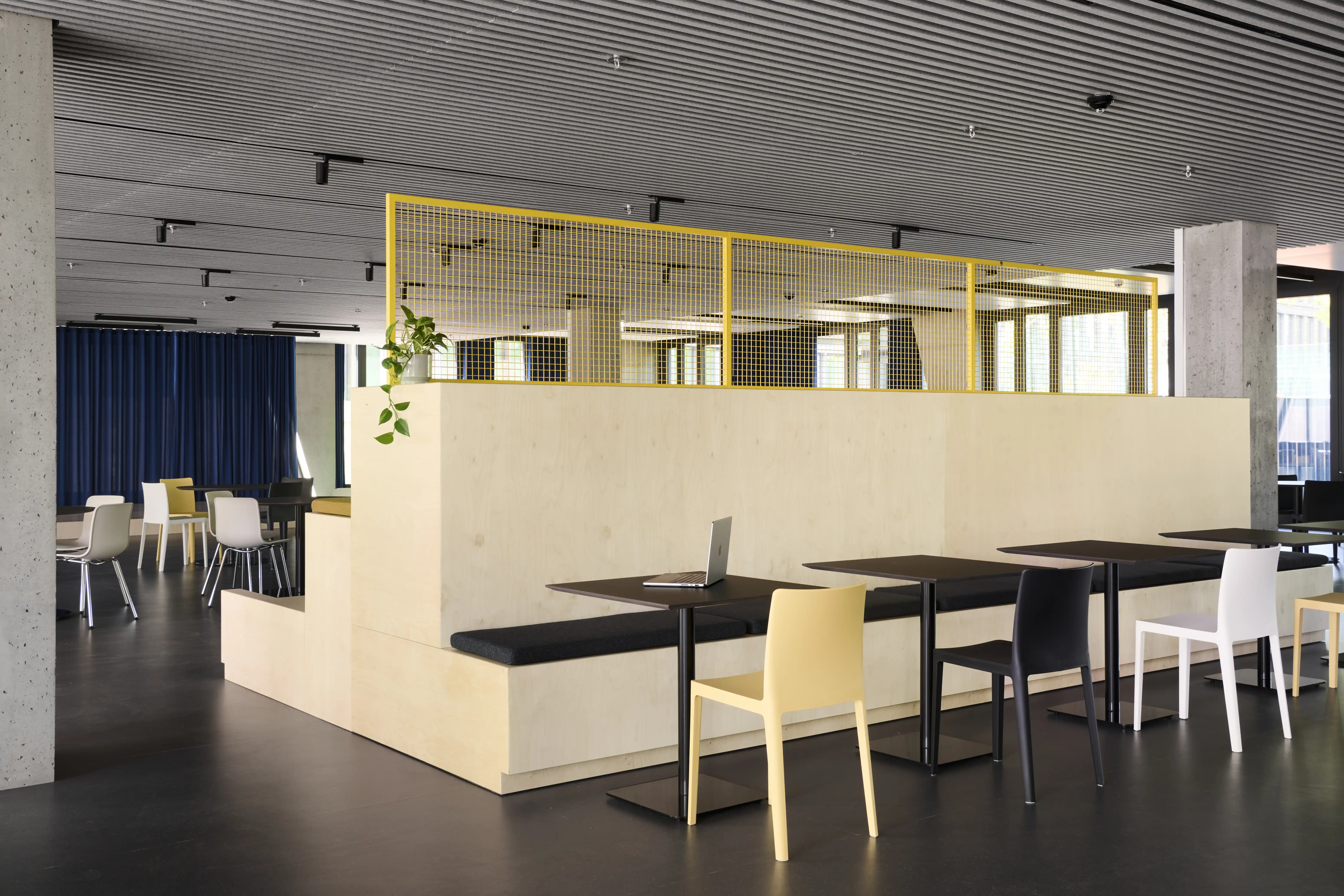 Birch seating tribune at ENPAL with black bench cushions, black bistro tables with yellow, white and black chairs and a yellow wire-mesh railing under a ribbed ceiling.