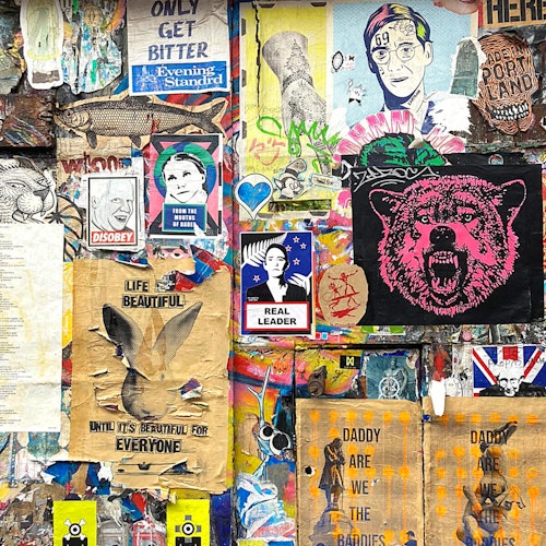 Isang makulay na pader na puno ng graffiti na puno ng iba't ibang poster, kabilang ang mga larawan ng umuungal na oso, mga pulitikal na mukha, at sining na batay sa teksto.