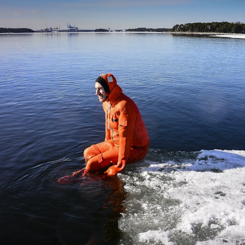 Person in an orange survival suit sits on the edge of an ice-covered shore, with a vast, calm body of water and distant trees in the background.