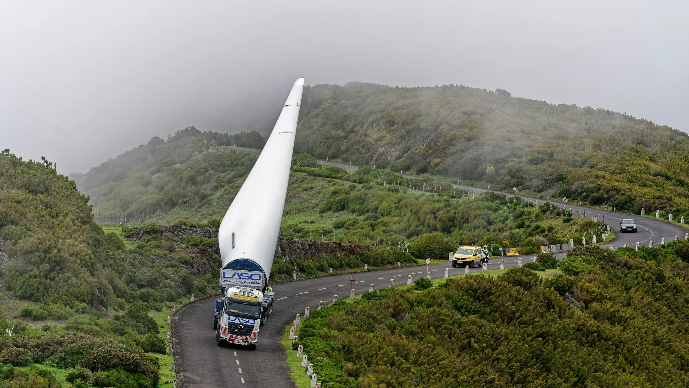 Truck hauling a long wind turbine blade along a winding road through green hills