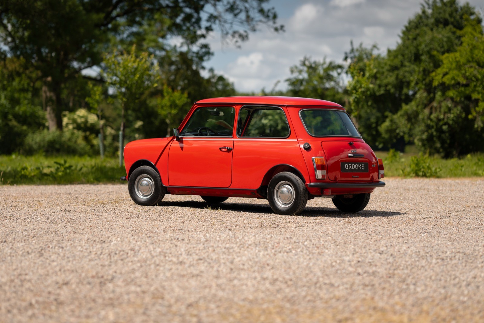A vintage red car parked on a gravel surface, surrounded by greenery and blue sky with clouds.