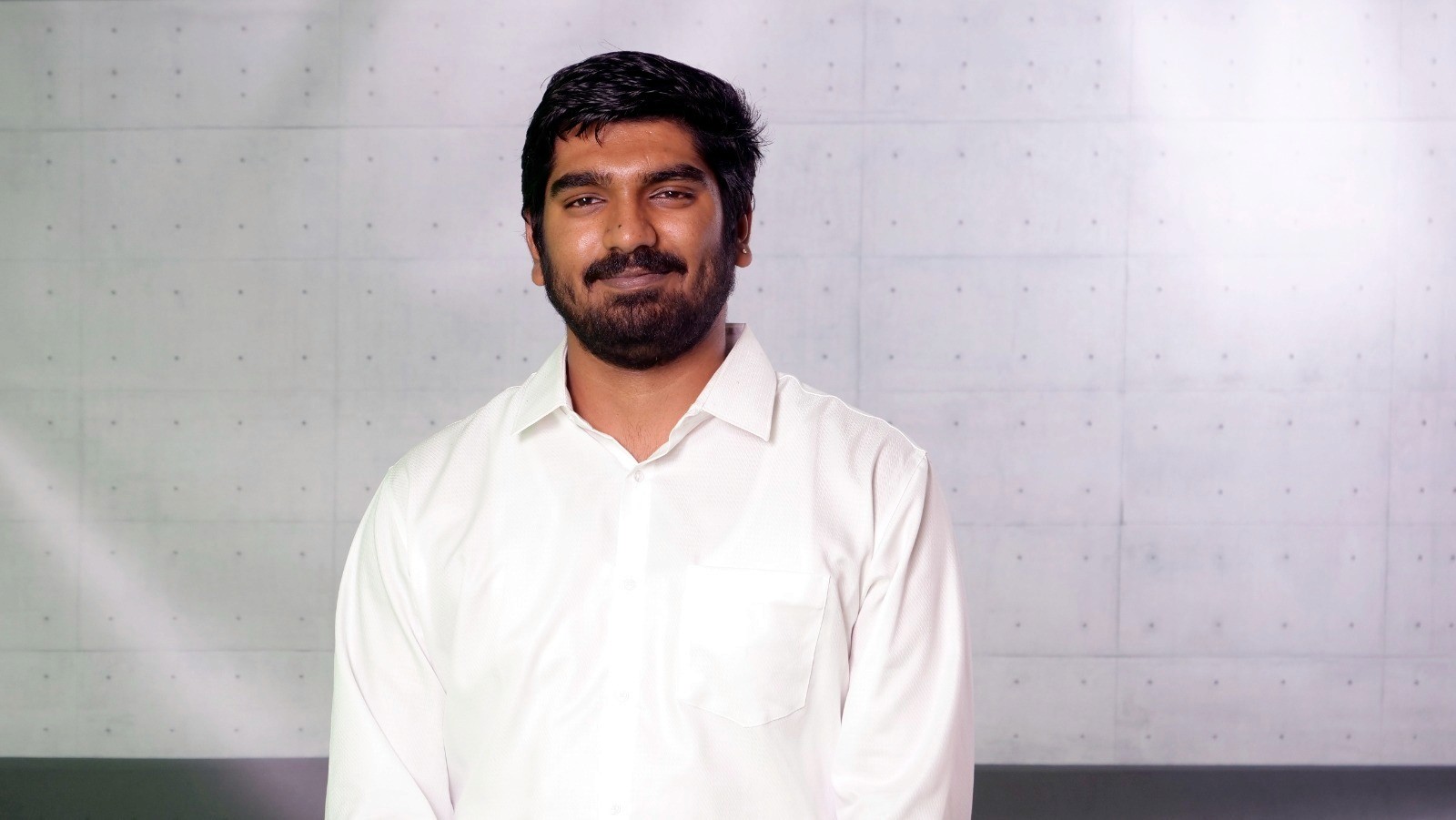 A smiling person with curly hair sits at a desk, looking at a laptop against a dark background.