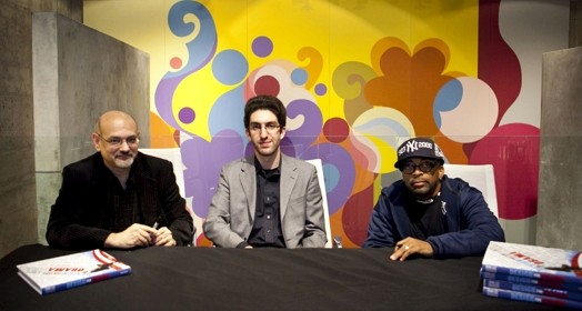 A photo showing three men sitting at a table with books on it and behind them some colorful wall art.