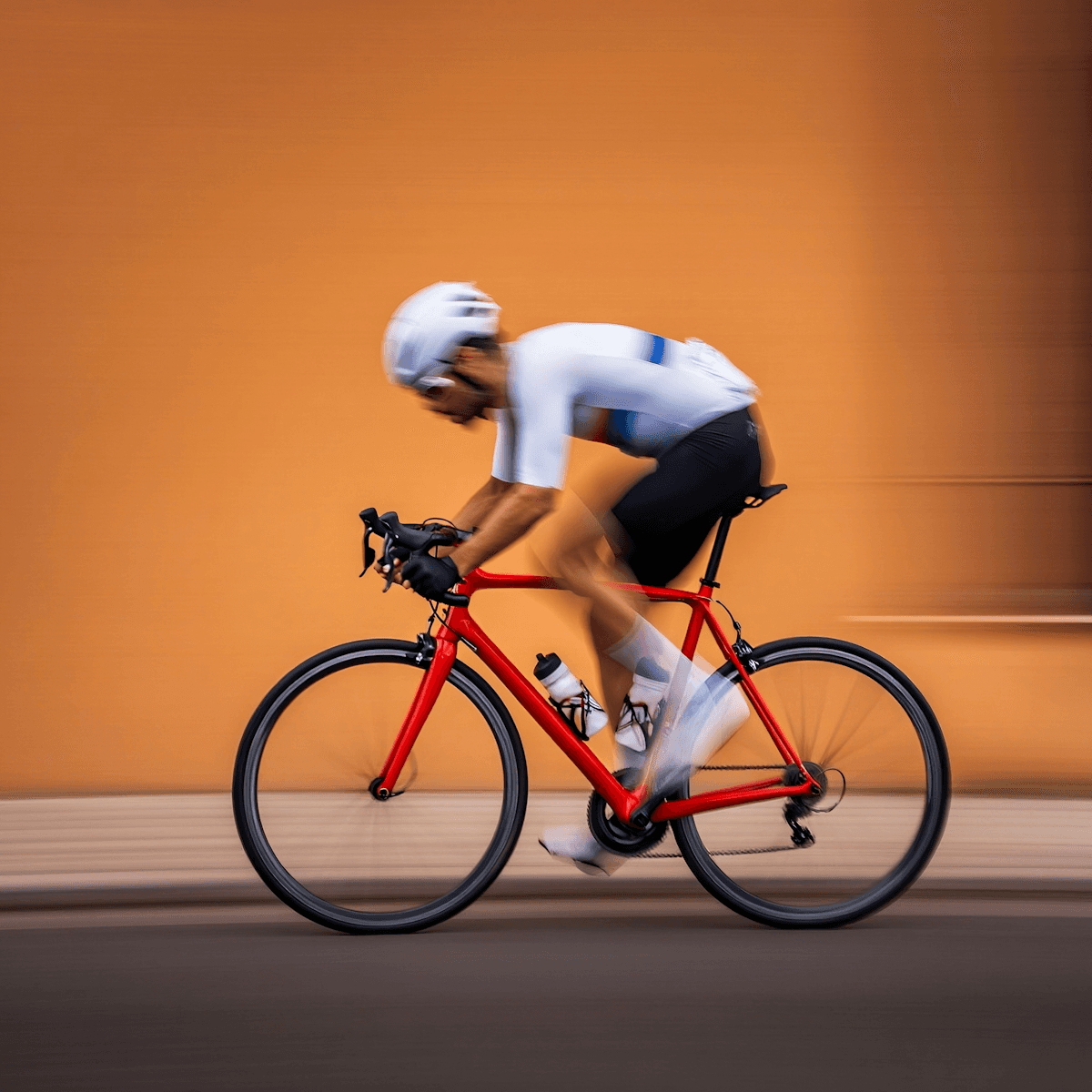 Cyclist wearing a white helmet and jersey rides a red bike against a blurred orange wall, conveying speed and motion.