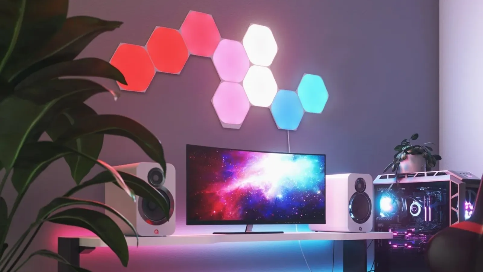 A modern gaming setup featuring a curved monitor displaying a colourful galaxy wallpaper, accompanied by sleek white speakers, illuminated hexagonal wall lights with a gradient of pink, red, and blue, and a custom-built gaming PC with vibrant RGB lighting, all set on a minimalist desk beside a lush green plant.