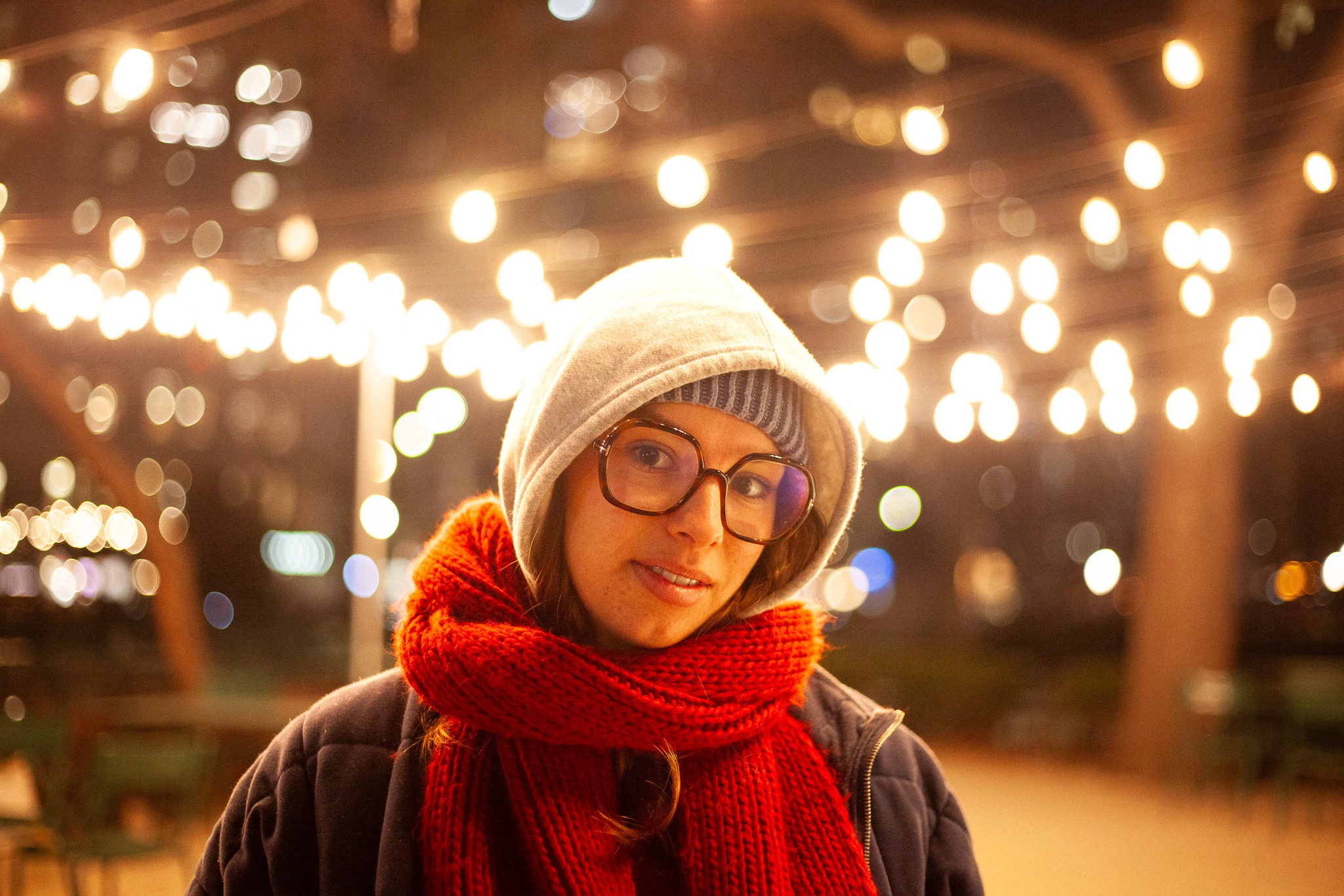 A person in a hoodie and red scarf stands in front of softly glowing string lights at night.