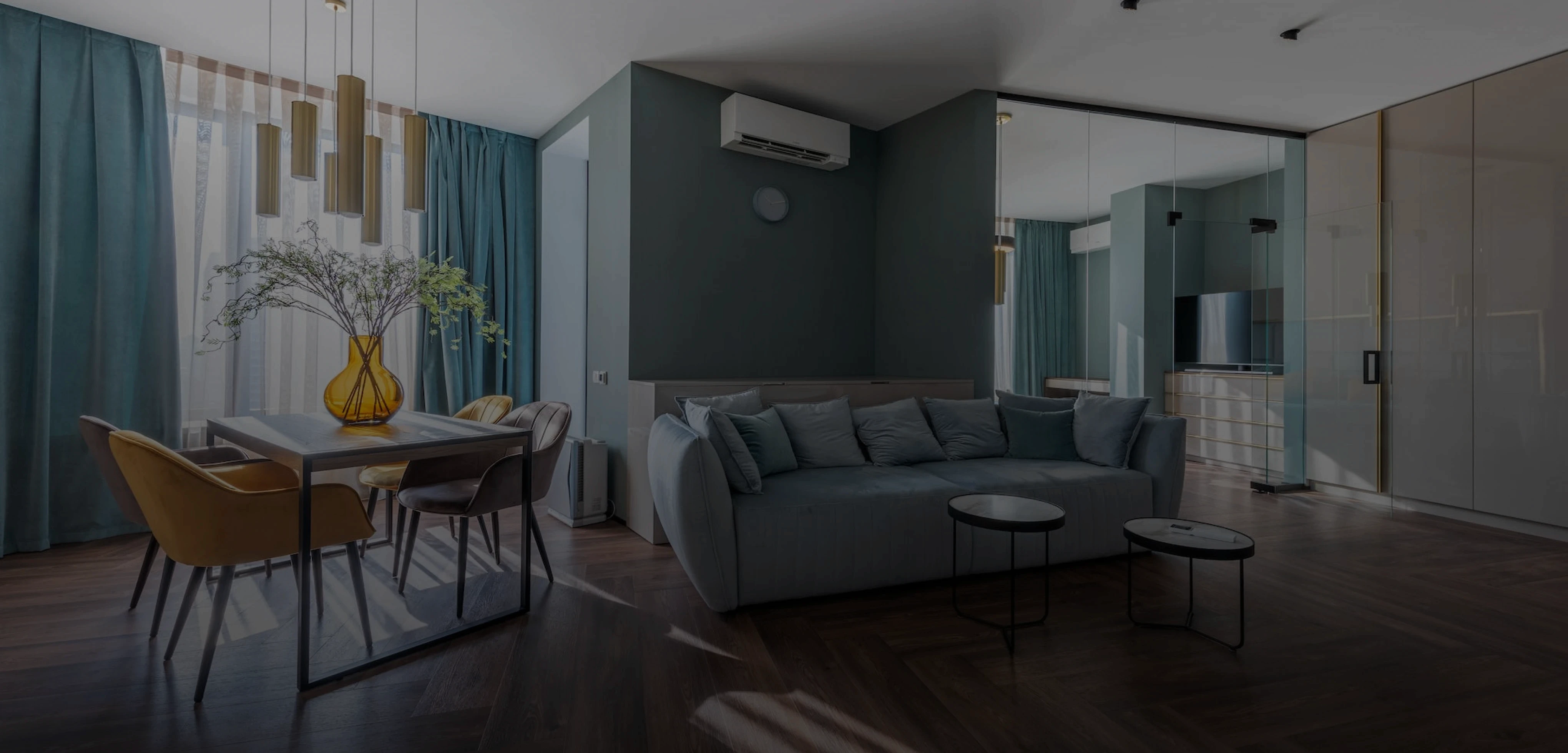 imagem da sala de uma casa elegante projetada por arquitetos. Imagem tem um sombreamento escuro para gerar contraste com o texto da seção. A sala apresenta cores pastéis em tons de azul, cinza e bege.