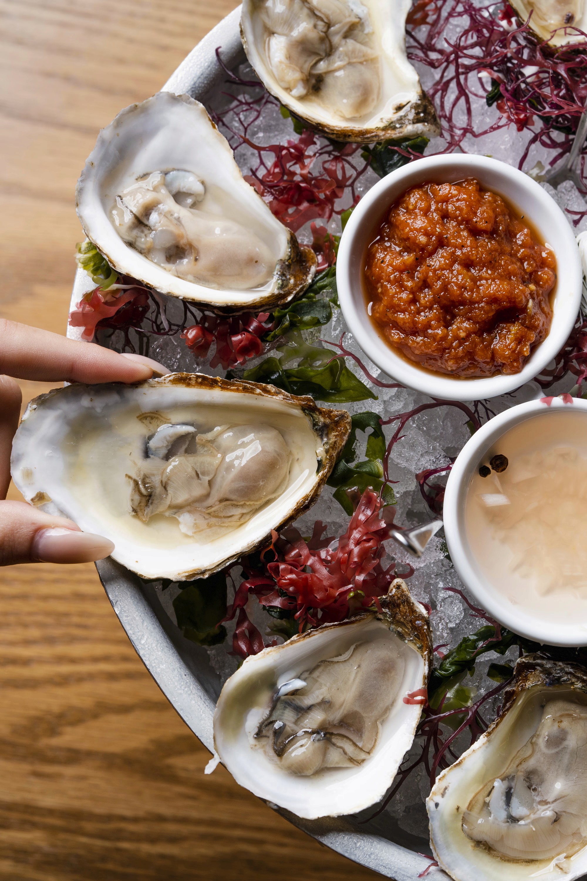 A wooden platter featuring oysters, sauces, and garnishes, with a hand reaching for an oyster.