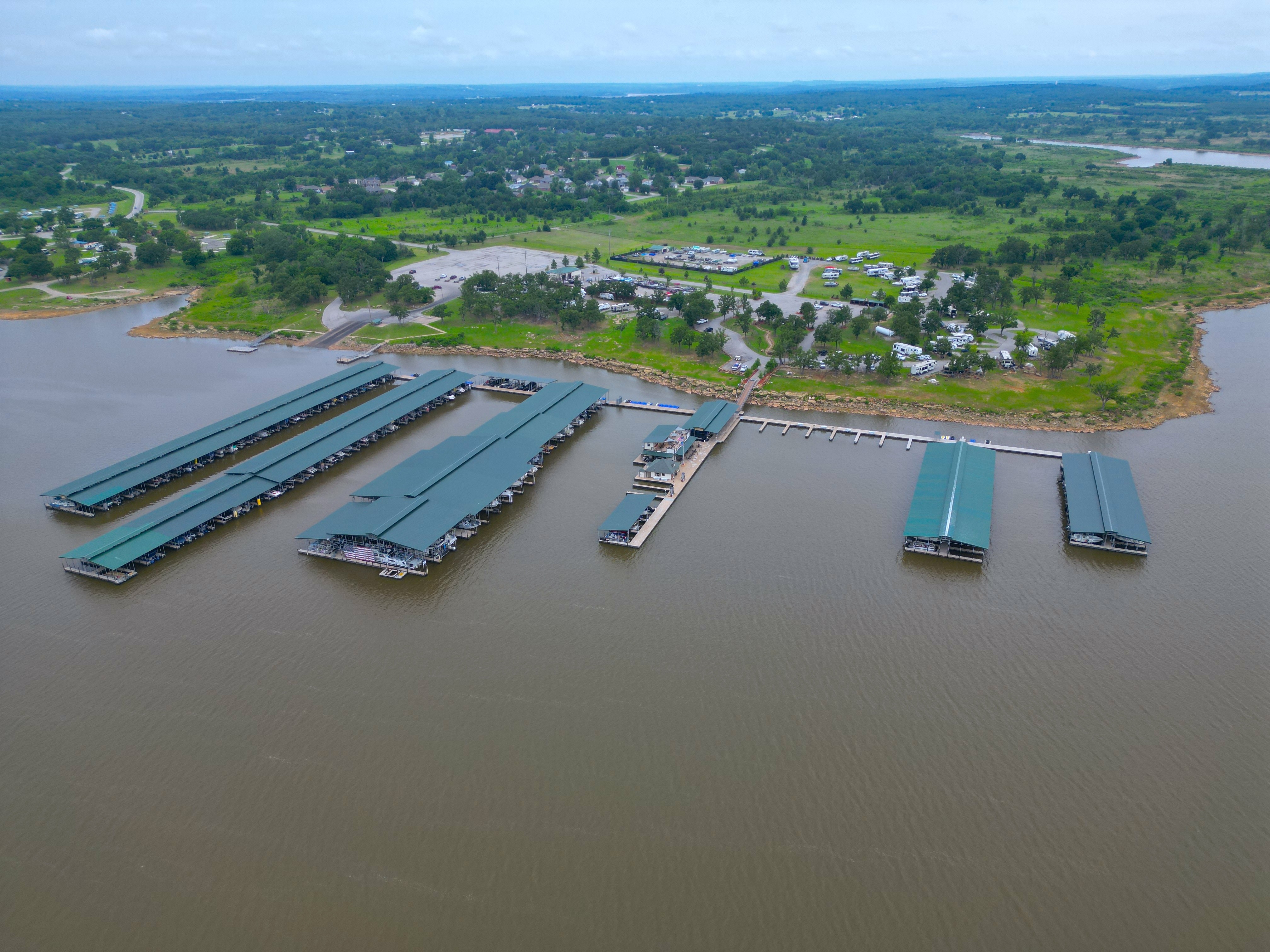Aerial view of a large keystone harbor marina on a calm, expansive keystone lake, featuring several long docks with green-roofed boat slips, surrounded by lush greenery and distant land.