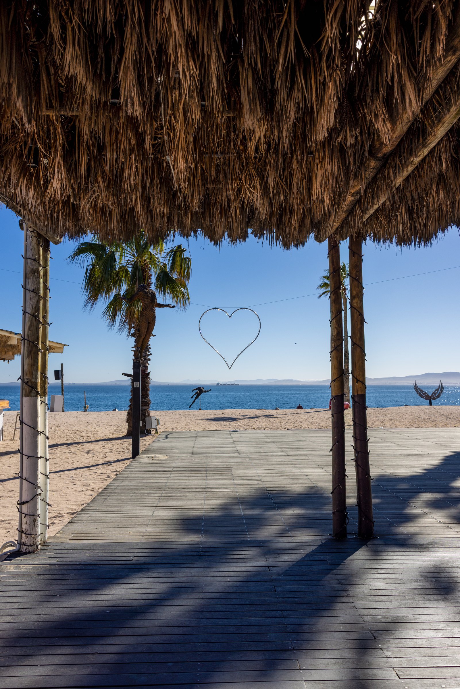 Palm-thatch walkway with fairy-light posts and a wire heart sculpture, leading out to the beach and sea