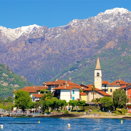 A picturesque lakeside village with colorful houses, a clock tower, and snow-capped mountains in the background.