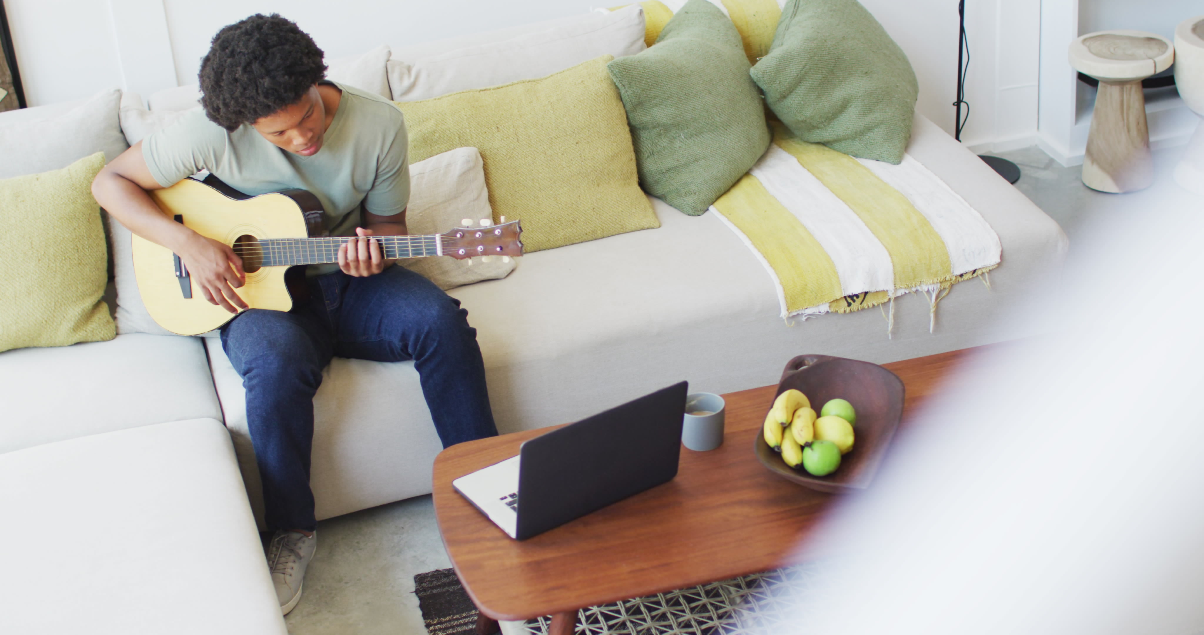 Young man sits on comfortable couch strumming guitar, focusing on laptop nearby.