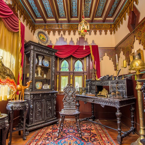 Intricately decorated room with ornate wooden furniture, colorful patterned rug, decorative objects, and rich red and gold drapery.