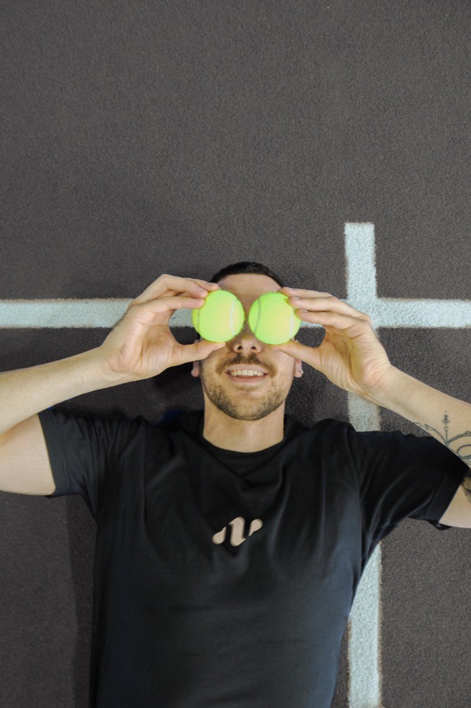 Padelspieler liegt lachend auf dem Court und hält zwei gelbe Bälle vor die Augen – ein verspielter Moment, der die humorvolle Seite von Padelnero zeigt.