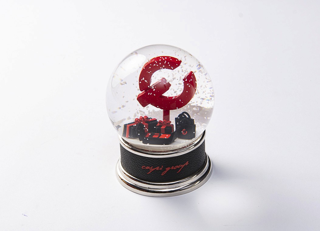 A close-up view of a snow globe featuring a large red "C" logo and miniature gift boxes surrounded by artificial snow.