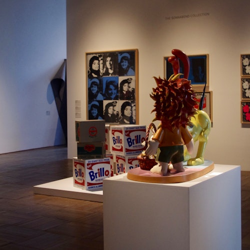 一处艺术画廊场景：摆放着 Brillo 盒子，底座上陈列着卡通风格的雕塑，墙上挂着装裱好的波普艺术肖像画。