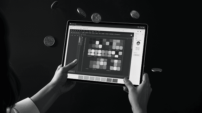 A black and white digital photograph of a person holding a tablet displaying a design or data visualization tool. The screen shows a grid of variously shaded squares with interface elements on the sides. The background is dark, with several coins appearing to be floating or flipping in mid-air, adding a surreal effect to the composition.